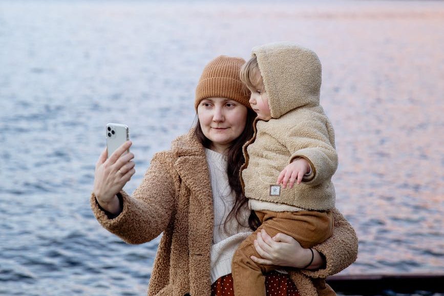 calm woman carrying adorable child on hands and taking selfie on smartphone near river and mountains